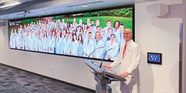 Surgical education center with a white coated medical professor standing at a podium. Next to him is a large videowall display with a photo of dozens of medical professors in white coats