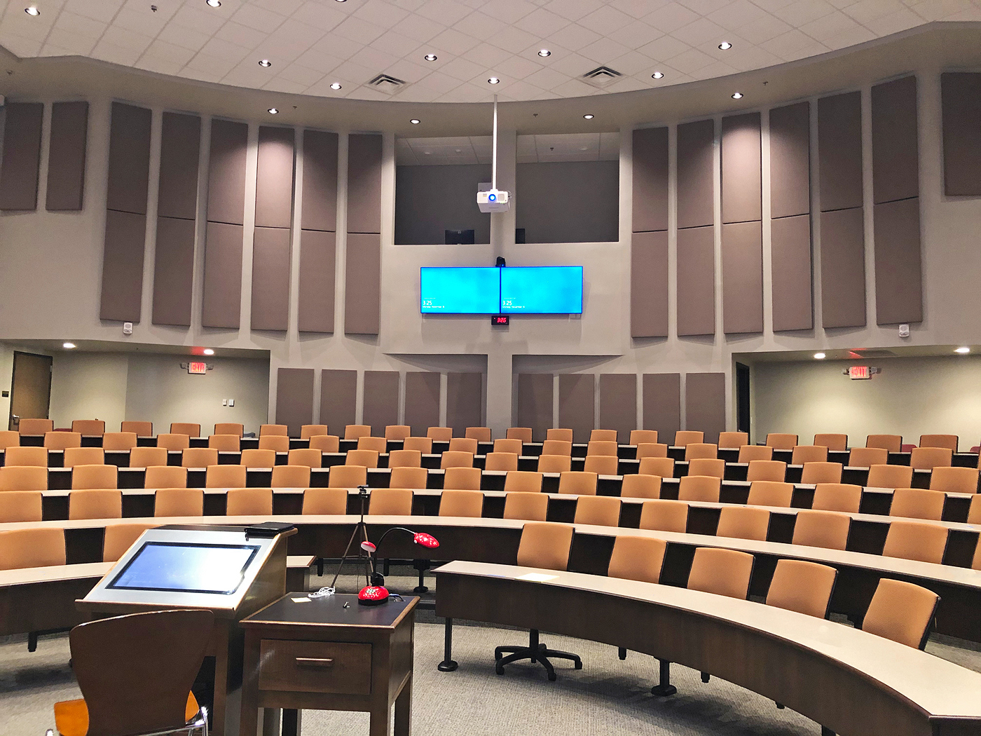 Lecture Hall viewed from presenter's position on-stage. The presenter podium in the foreground has a touchscreen for annotation and a document camera. Two flat panel display confidence monitors and a PTZ camera face the stage. Windows allow viewing from the second-floor mezzanine.