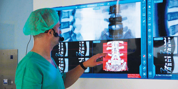 Surgeon in scrubs wearing a microphone and pointing to large touchscreen display showing spinal anatomy