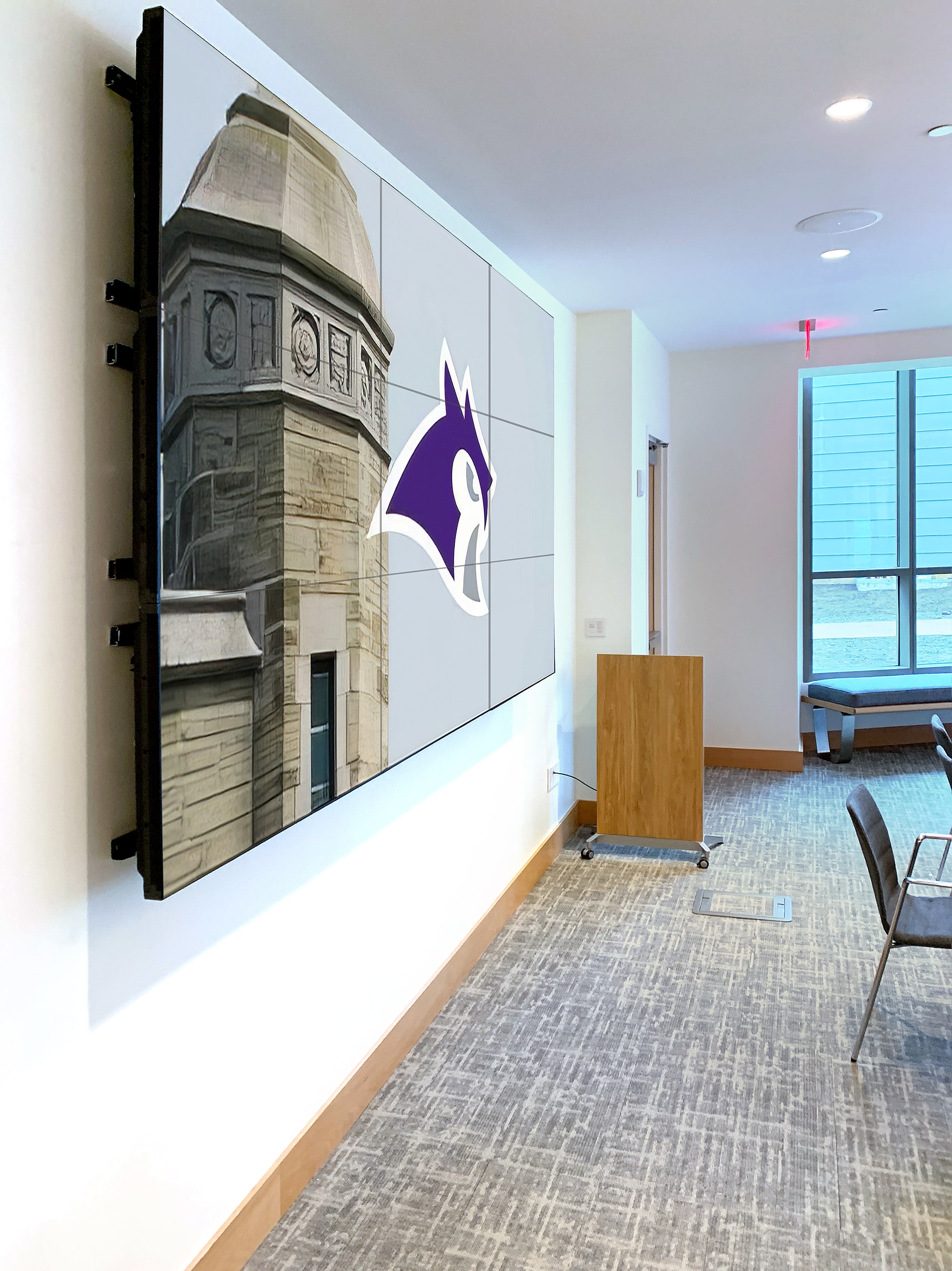 Cuando una reunión del personal o un seminario se celebra en esta sala de reuniones de Lowell House, el presentador envía contenido al videowall de 3x3 en la parte delantera de la sala. Los decodificadores escaladores NAV con la tecnología WindoWall™ de Extron hacen funcionar el videowall.