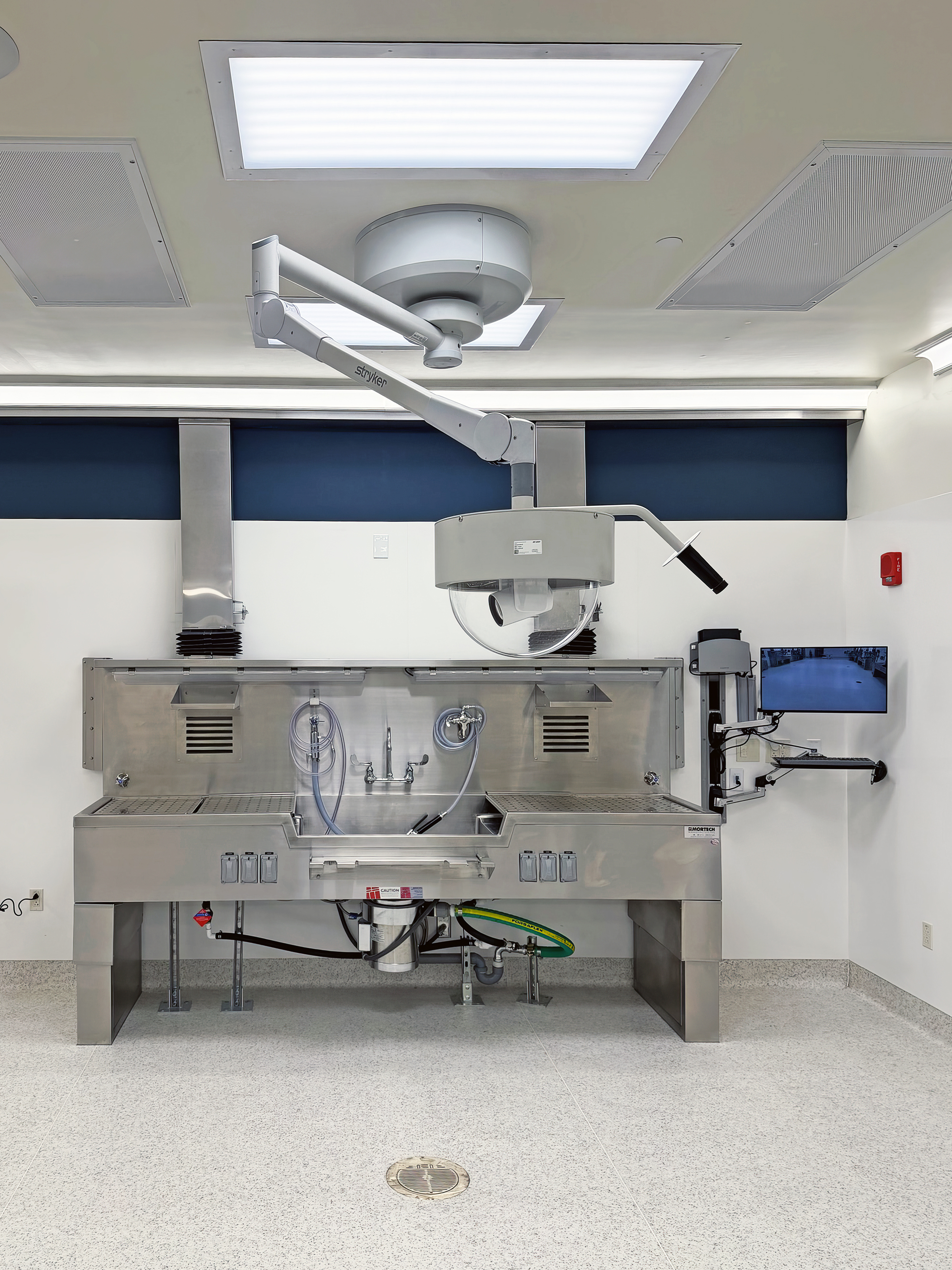 The autopsy room includes bright lighting, stainless steel work surfaces, and space for staff movement. Cameras and AV connections support live viewing and recording.