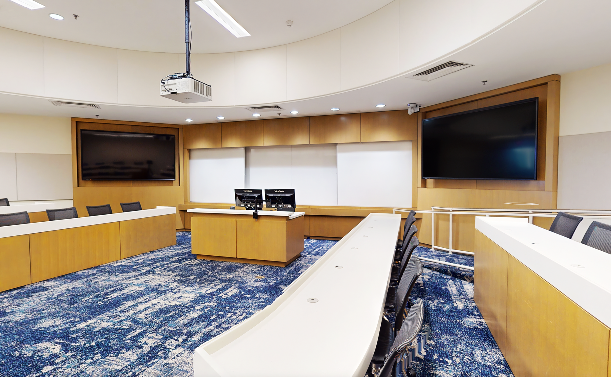 Vue de face de la salle de classe Glaxo montrant le bureau de l'enseignant équipé de deux écrans plats, un projecteur avec écran de projection motorisé qui s'abaisse sur les tableaux blancs, et une caméra PTZ à commande manuelle installée au plafond. 