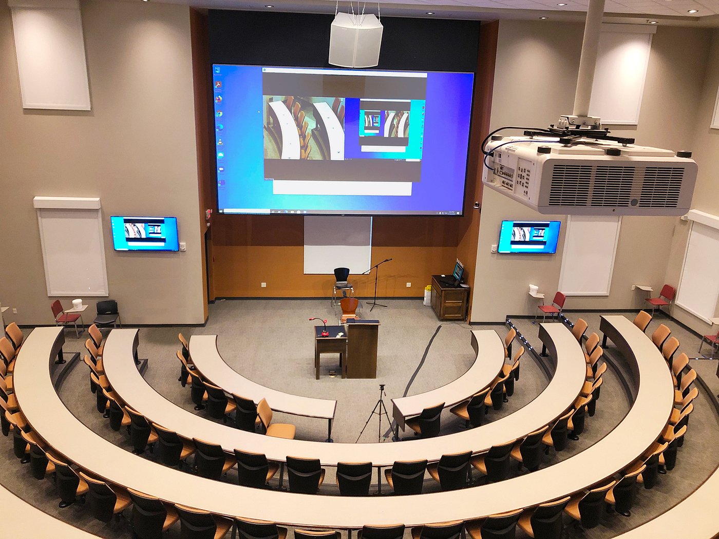 Lecture Hall viewed looking toward stage. The presenter podium is at center stage. The AV operator desk is stage left. The projector is seen in the foreground. The projection screen is flanked by two flat panel displays. A PTZ camera facing the audience is mounted to the wall at the lower right corner of the projection screen.