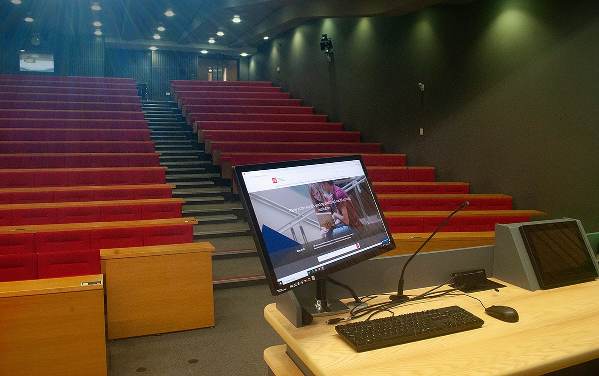 Un moniteur sur bras articulé et un clavier offrent aux présentateurs un accès direct au contenu numérique, entouré des sièges en gradins de l'amphithéâtre.