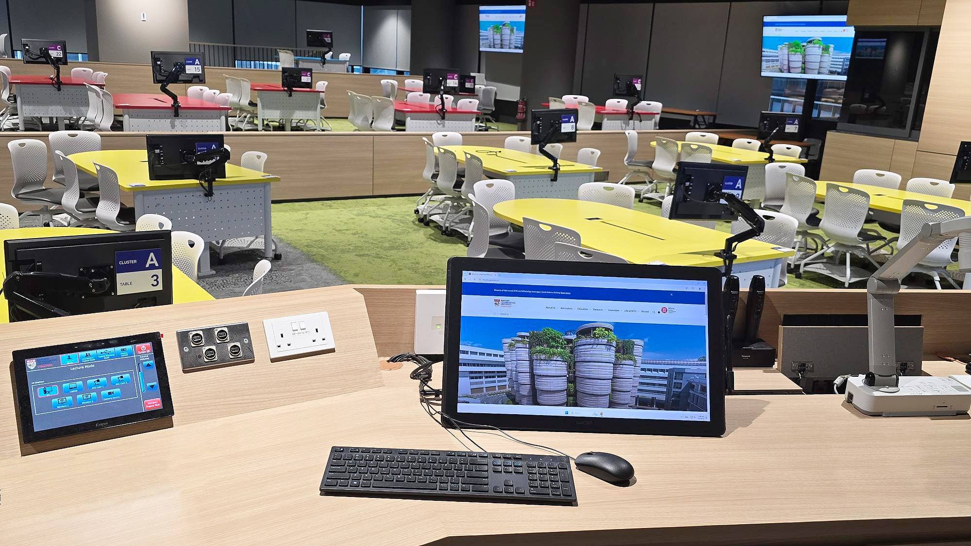 Vue depuis le bureau de l'enseignant de la salle de collaboration 1 montrant l'écran tactile intuitif Extron, intégré de manière optimale à côté des outils de bureau standard.