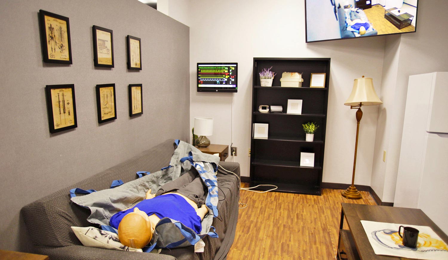 High fidelity medical mannequin lies supine on a sofa in the living room of the Emergency Medical Services simulation suite. The mannequin’s medical symptoms are controlled by instructors in an adjacent control room. Vital signs are displayed on the computer monitor on the far wall and images captured by a PTZ camera in the room appear on the 55” flat panel display in the upper corner of the far wall.