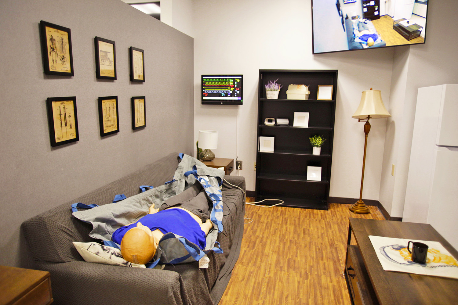 High fidelity medical mannequin lies supine on a sofa in the living room of the Emergency Medical Services simulation suite. The mannequin’s medical symptoms are controlled by instructors in an adjacent control room. Vital signs are displayed on the computer monitor on the far wall and images captured by a PTZ camera in the room appear on the 55” flat panel display in the upper corner of the far wall.