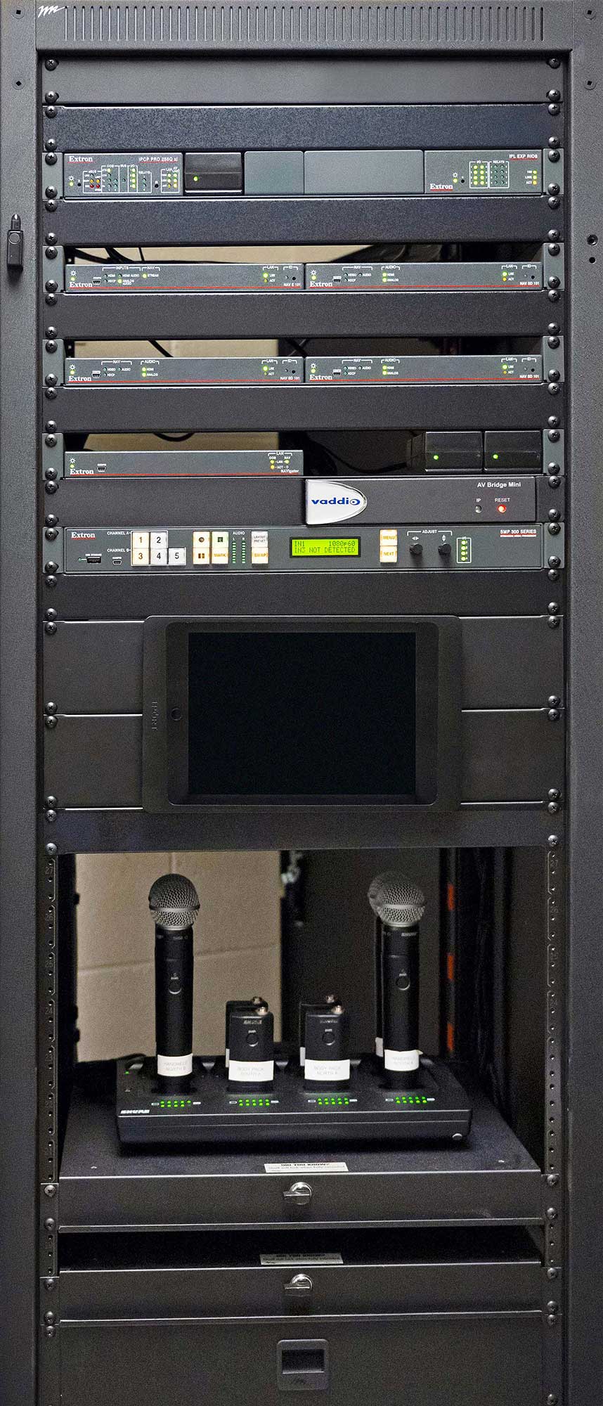AV-Rack. Von oben nach unten: IPCP Pro-Steuerungsprozessoren, NAV Pro-Kodierer und -Decoder, NAVigator, SMP 352-Prozessor für Medienstreaming sowie iPad und kabellose Mikrofone auf Ladestationen.