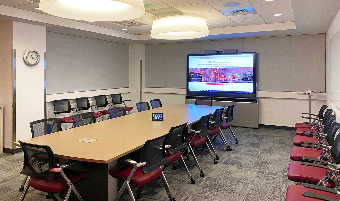 An empty medical conference room, with seating along the walls as well as around the table, in which a touchpanel is being used to control the large wall display.