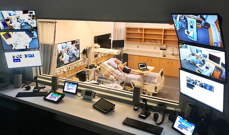 Una sala de control en primer plano que mira a través de una ventana a un laboratorio de simulación de destrezas médicas.