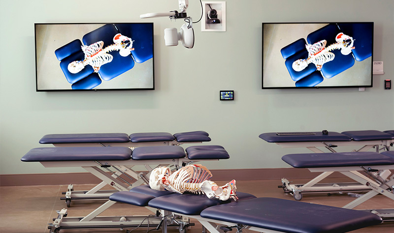 Un laboratorio de destrezas en una escuela de quiropráctica con mesas ajustables, cámaras suspendidas y dispositivos de visualización.